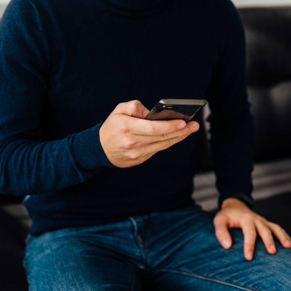 Iemand in een donkerblauwe trui en spijkerbroek zit op een leren bank en kijkt op de smartphone in zijn hand.